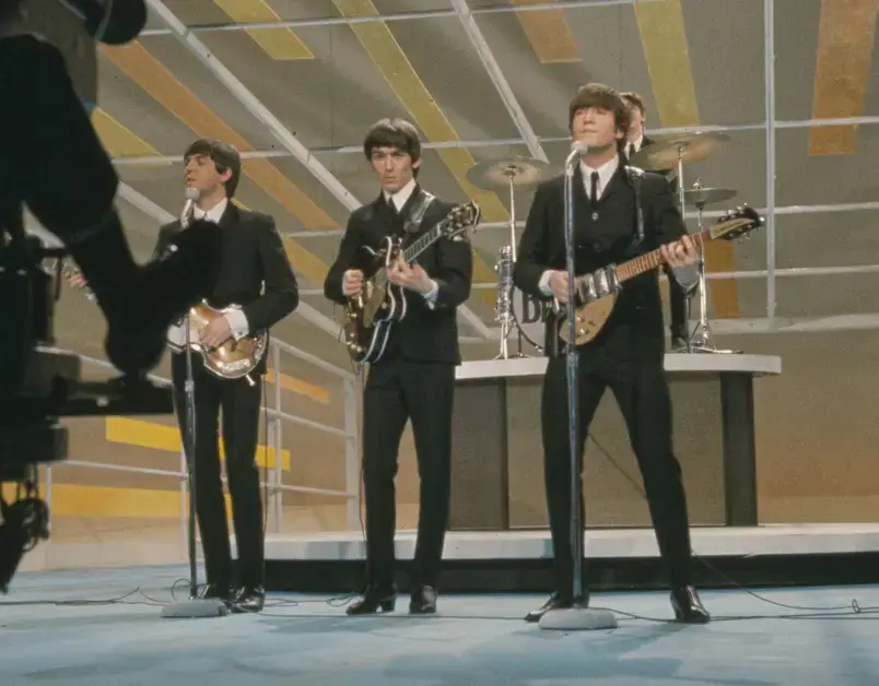 Os Beatles tocando em um programa de TV nos anos 60, vestindo ternos escuros e gravatas.