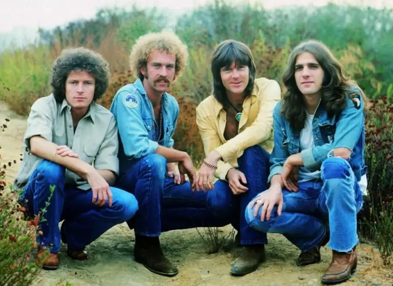 A imagem mostra membros da banda de rock Eagles, que formaram a formação original. Eles são, da esquerda para a direita, Don Henley, Bernie Leadon, Randy Meisner e Glenn Frey. Eles estão agachados numa paisagem rural. A imagem serve para ilustrar o artigo Eagles: Além de Hotel California.