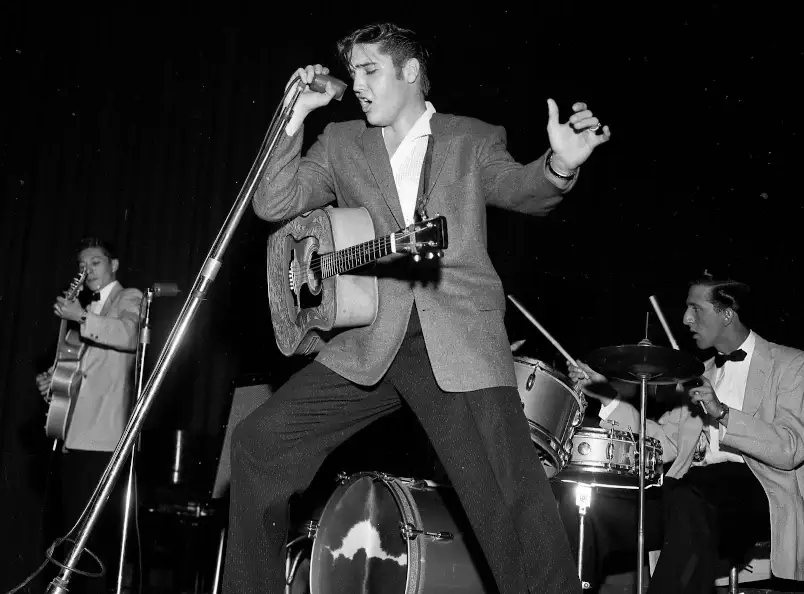 Elvis Presley cantando em um palco nos anos 50, com um violão e segurando um microfone